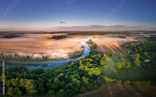 Fototapeta Naklejka Na Ścianę i Meble -  Light rays trough thick fog over a river in a misty summer morning. Rural landscape covered with pine forest and farm land.