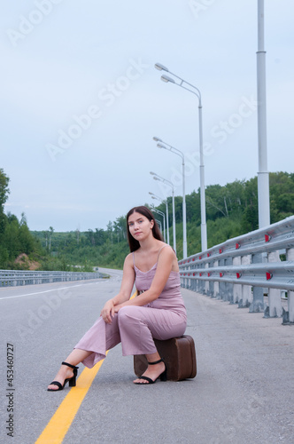 A girl is sitting on a suitcase on the side of an empty road waiting for a car