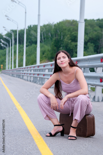 A girl is sitting on a suitcase on the side of an empty road waiting for a car © Beliakina Ekaterina