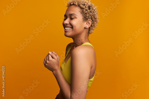 Horizontal studio shot of cute, chic young African female model in lovely yellow crop top posing against orange wall half-turned with cheerful face expression, eyes closed fists pressed together