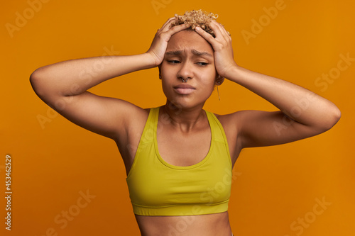 Migraine. Good-looking young dark-skinned woman looking sad and depressed, holding blonde curly head with hands, suffering from stress, headache, feeling sick, wearing yellow fitness clothes