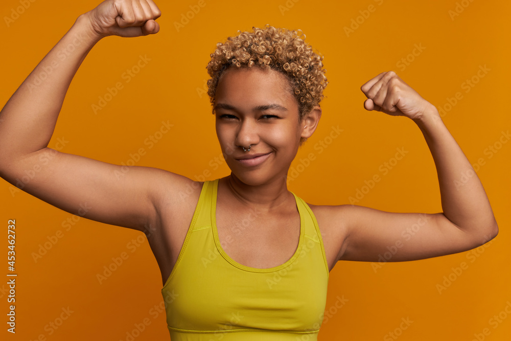 Close-up studio picture of sporty ethnicity female with stylish haircut showing strength, power, muscles, putting arms up, smiling, feeling proud of herself. Fitness and healthy lifestyle