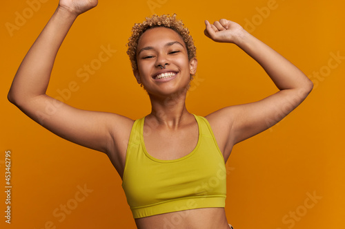 Picture of pretty cheerful young dark-skinned sporty female model with short blonde curls stretching muscles, doing warm up before fitness training exercises, having satisfied toothy smile