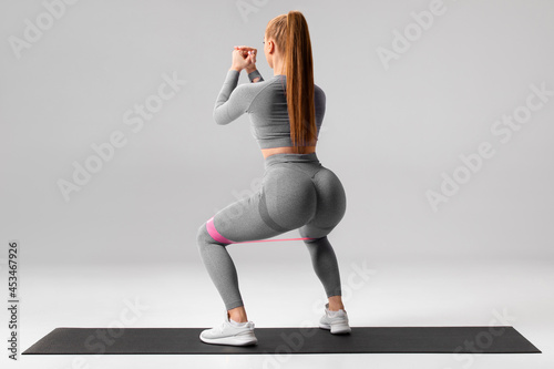Athletic girl doing squats exercise for glute with resistance band on gray background. Fitness woman working out