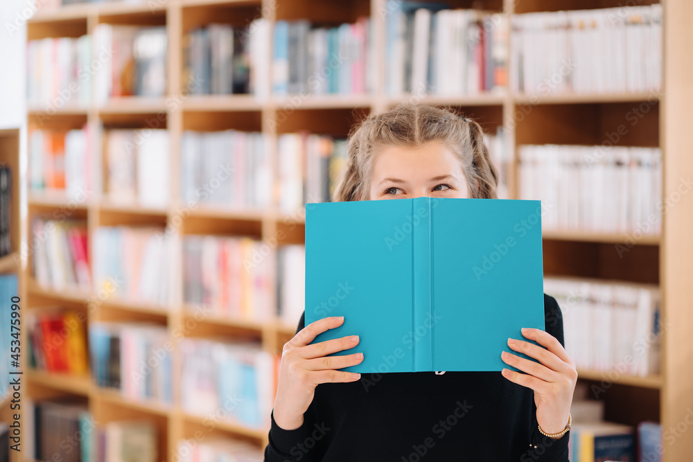 Portrait of a pretty young girl hiding behind an open blue book and looking away over bookshelf background Portrait of a pretty young girl hiding behind an open blue book and looking away over bookshelf background