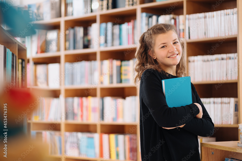 Happy tenage girl or smiling student holding a blue book wih copy space among many books in library - People, knowledge, education and school concept Happy tenage girl or smiling student holding a blue book wih copy space among many books in library - People, knowledge, education and school concept