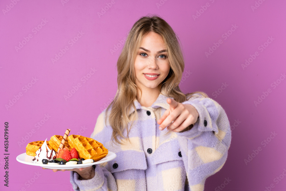 Teenager Russian girl holding waffles isolated on purple background points finger at you with a confident expression