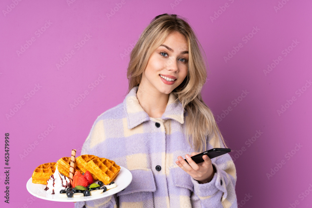 Teenager Russian girl holding waffles isolated on purple background holding coffee to take away and a mobile Teenager Russian girl holding waffles isolated on purple background holding coffee to take away and a mobile