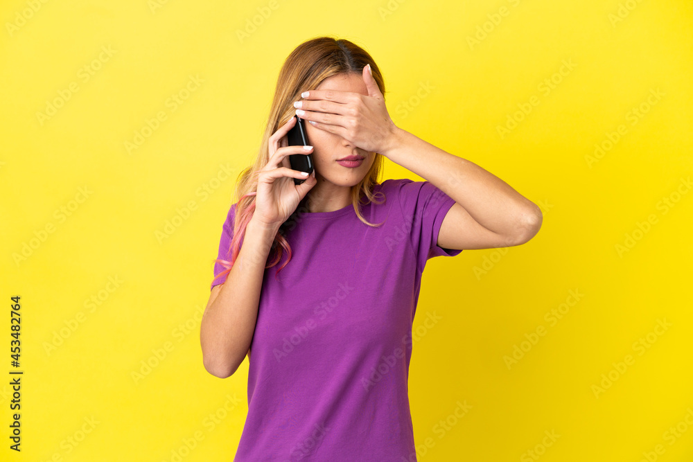 Young woman using mobile phone over isolated yellow background covering eyes by hands. Do not want to see something Young woman using mobile phone over isolated yellow background covering eyes by hands. Do not want to see something
