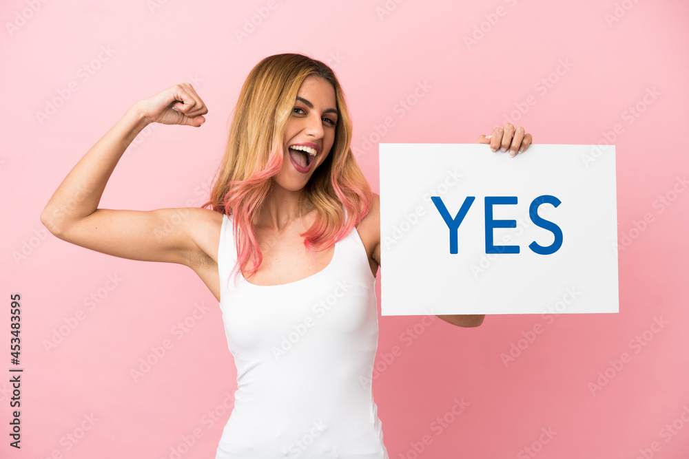 Young woman over isolated pink background holding a placard with text YES doing strong gesture Young woman over isolated pink background holding a placard with text YES doing strong gesture