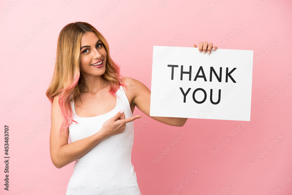 Young woman over isolated pink background holding a placard with text THANK YOU and pointing it Young woman over isolated pink background holding a placard with text THANK YOU and pointing it
