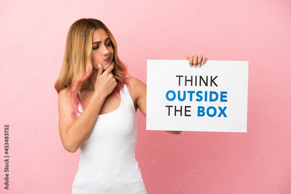 Young woman over isolated pink background holding a placard with text Think Outside The Box and thinking Young woman over isolated pink background holding a placard with text Think Outside The Box and thinking