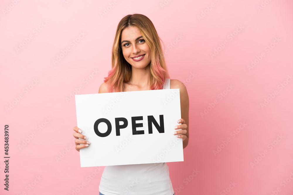 Young woman over isolated pink background holding a placard with text OPEN with happy expression Young woman over isolated pink background holding a placard with text OPEN with happy expression