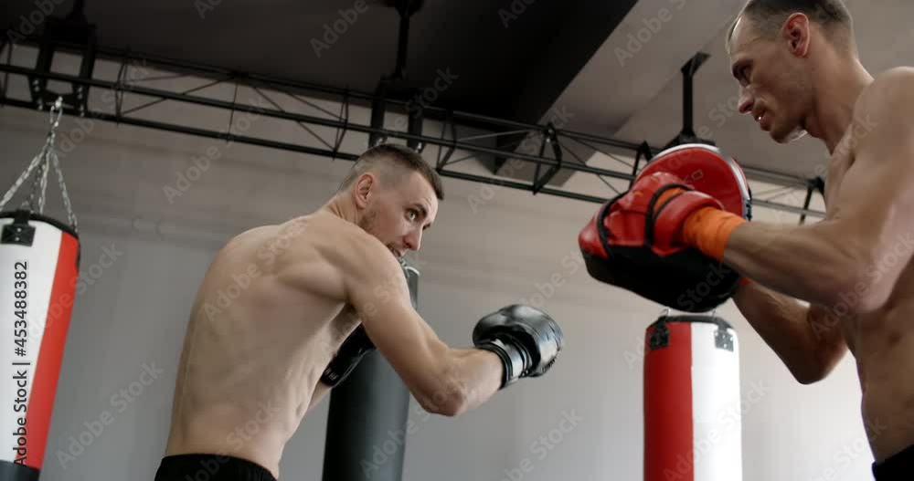 Low angle shot: fighter in boxer gloves is practising his strikes with trainer in slow motion in the gym, kickboxers are training, 4k 120fps Prores HQ Low angle shot: fighter in boxer gloves is practising his strikes with trainer in slow motion in the gym, kickboxers are training, 4k 120fps Prores HQ