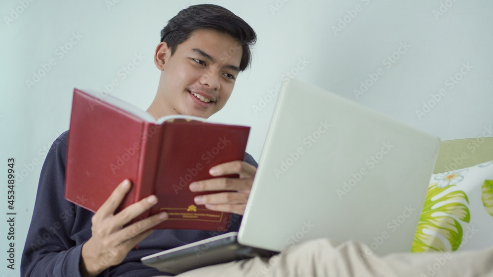 young asian man holding book happily while looking at laptop screen at home
