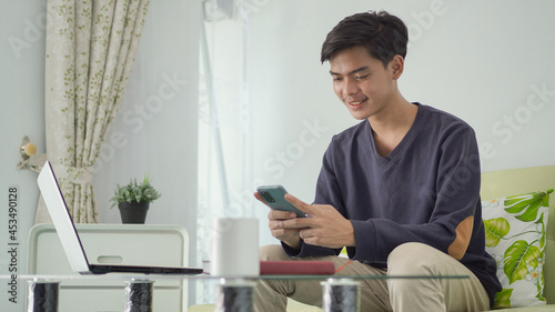young asian man playing smartphone in front of laptop screen at home