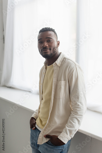 african american man in shirt standing with hands in pockets near window sill