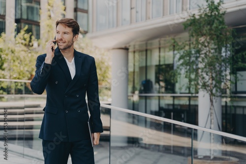 Young man office worker in suit talking on mobile phone while walking outside of business center