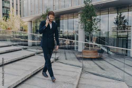 Happy caucasian businessman talking on smartphone while walking along office center © VK Studio