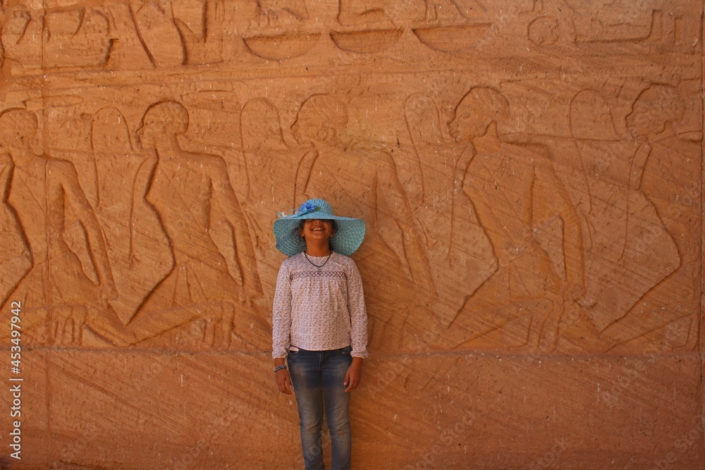 A young girl with a hat standing in front of an ancient wall in Abou simbel temple featuring prisoners in Aswan in Egypt
