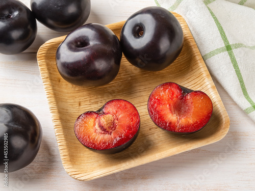 Black plums whole and cut in half on a square wooden plate over table. Ready to eat fresh ripe plums with red juicy pulp. Close-up of tasty sweet fruits. Vegetarian, vegan and vitamin healthy eating.