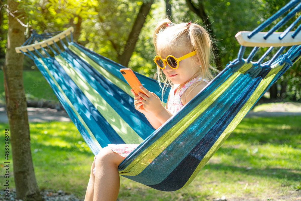 Cute little girl in a yellow sunglasses holding smartphone and resting on a hammock in the park Cute little girl in a yellow sunglasses holding smartphone and resting on a hammock in the park