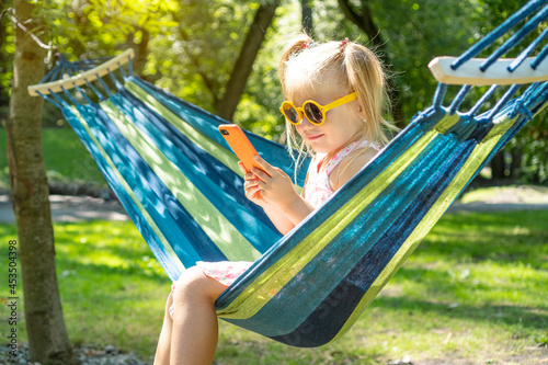Cute little girl in a yellow sunglasses holding smartphone and resting on a hammock in the park