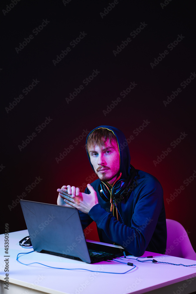 Hacker with a laptop sits at the table, dark background and neon light behind, head covered with a hood. Hacker with a laptop sits at the table, dark background and neon light behind, head covered with a hood.