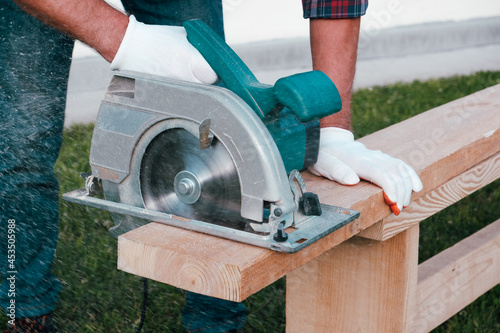 Working with a power tool. A builder is sawing a board at the construction site of a new house. Close-up on gloved hands holding a circular saw. photo