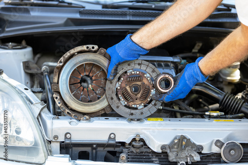 Automotive technician holding used car pressure plate, clutch disc and release bearing in front of the vehicle engine