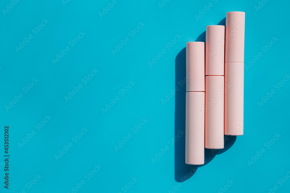 Fototapeta premium pink tubes of cosmetics on a blue background. Copy space. Closed tubes of lipstick, liquid lip gloss, mascara, eyeliner