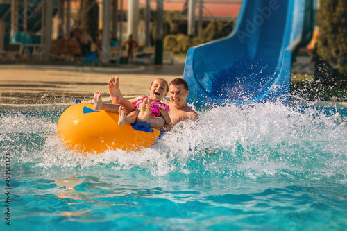 Summer holidays on a sunny day in an outdoor water park. A father and a child ride on a rubber circle from a slide into the water on a water attraction.