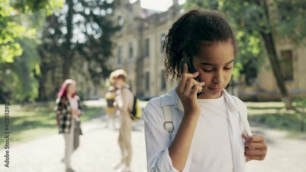 Sad school girl talking phone looking at happy friends hanging out after classes Sad school girl talking phone looking at happy friends hanging out after classes