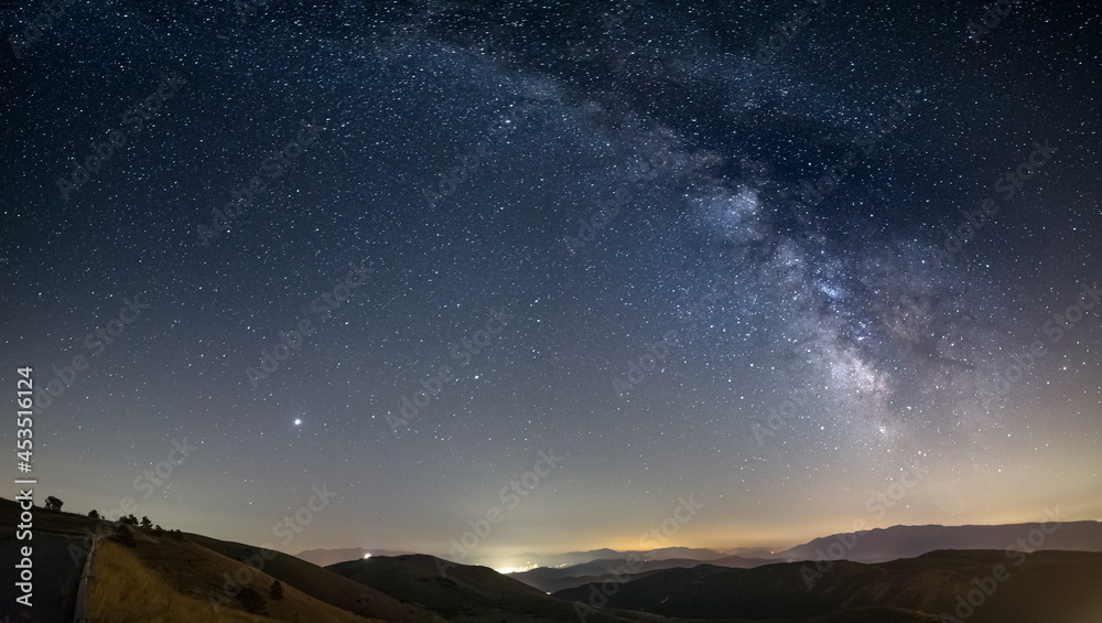 Panoramic night sky over Santo Stefano di Sessanio, Abruzzo and Rocca Calascio, Italy. The Milky Way galaxy arc and stars over illuminated village unique hills landscape. Jupiter planet visible.