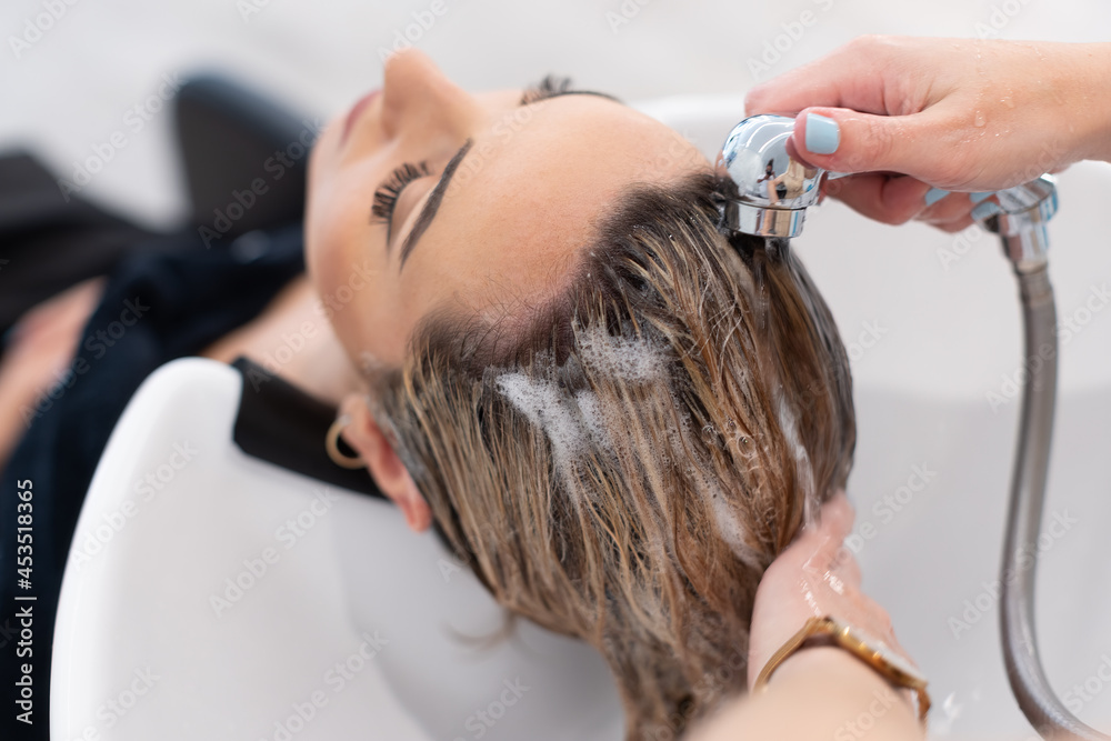 Fototapeta premium Hair stylist at work - hairdresser washing blond hair to the caucasian woman customer with eyes close. Selective Focus