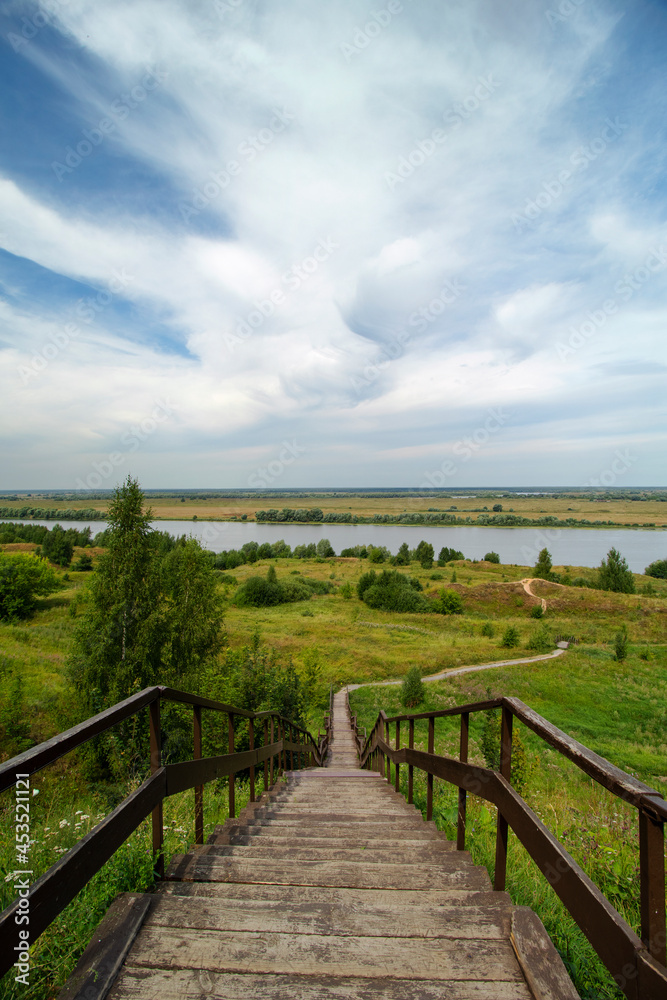 Vertical photo of a wooden staircase and descent from the mountain to the Oka River on a clear summer day. A winding path through the hills going to the river. The village of Konstantinovo.