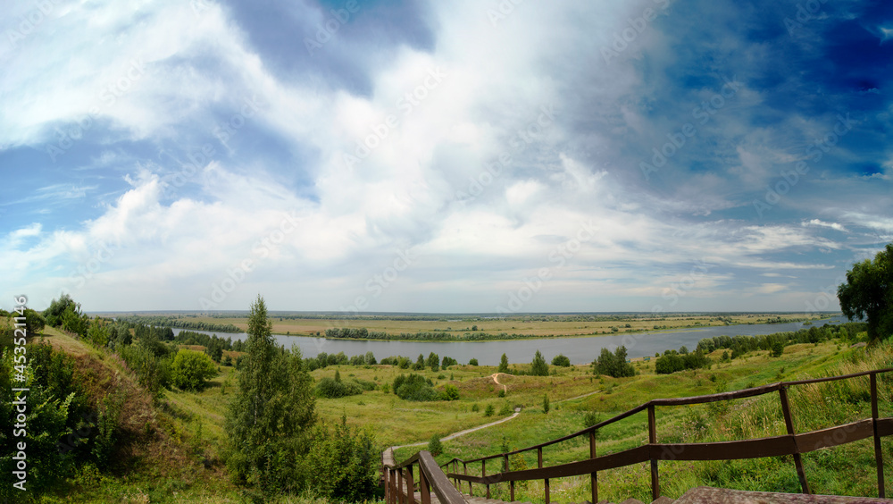 Panoramic photo of the descent from the mountain to the Oka River on a clear summer day. A winding path through the hills going to the river. The village of Konstantinovo.