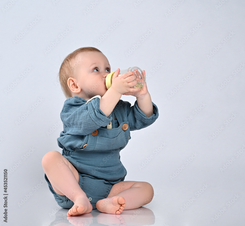 oddler toddler in a blue staple overalls, sitting on the floor barefoot and drinking water from a bottle with a pacifier. oddler toddler in a blue staple overalls, sitting on the floor barefoot and drinking water from a bottle with a pacifier.