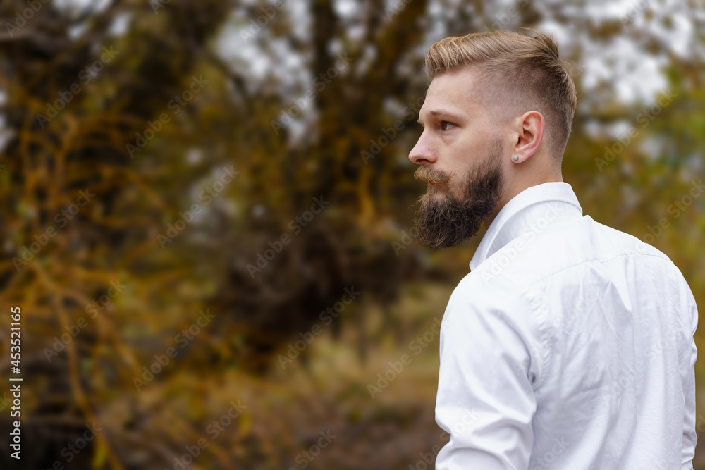 Young bearded man in white shirt walks on yellow autumn day in park. Spending time alone in nature. Calming atmosphere. Looks into the distance. Reflects on outdoor life
