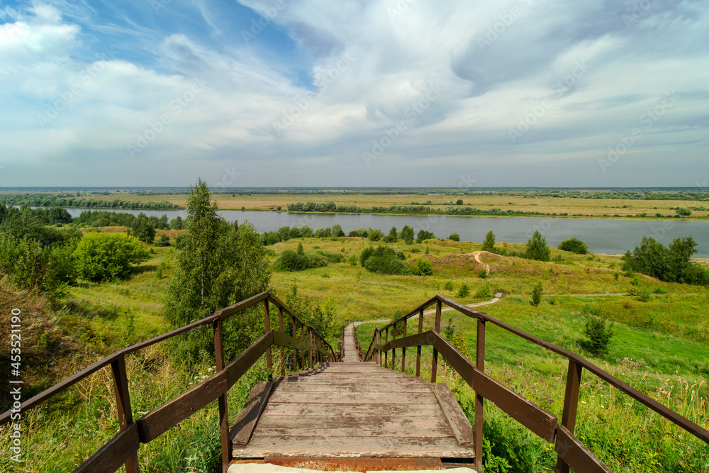 Horizontal photo of a wooden staircase and descent from the mountain to the Oka River on a clear summer day. A winding path through the hills going to the river. The village of Konstantinovo.