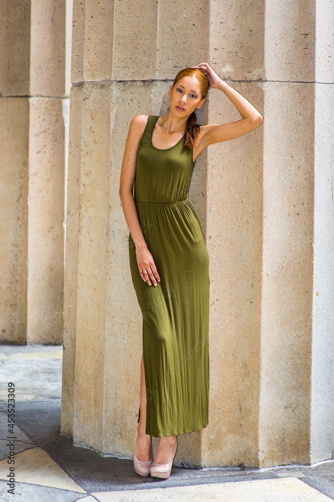 Dressing in a green Maxi Tank Dress, creamy high heels,  crossing legs, one hand touching her head, one hand resting on her thigh, a young fashion black girl is standing  by a column, relaxing.