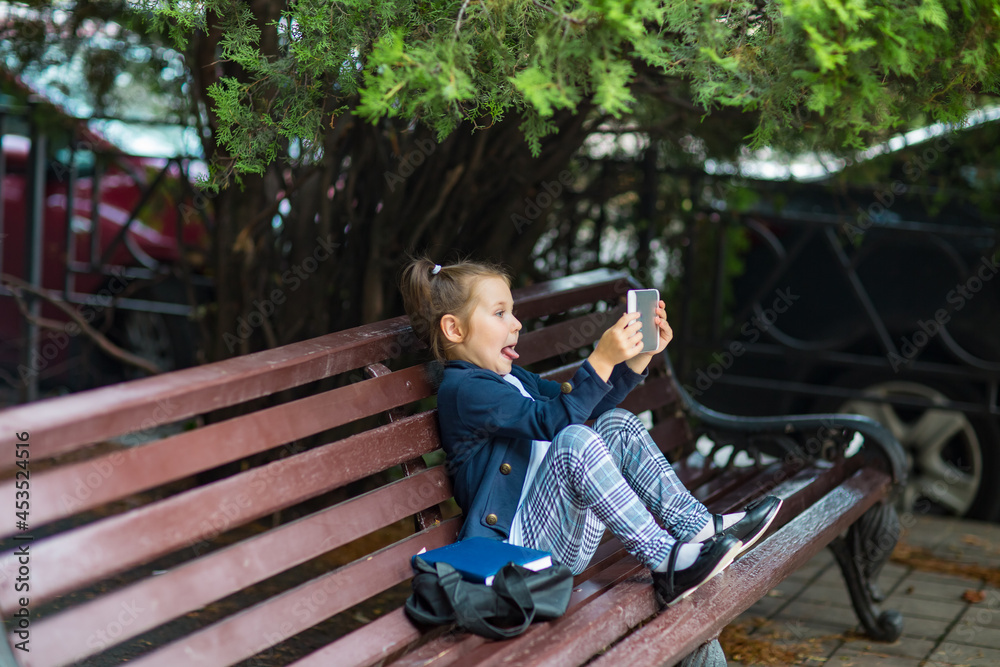 a little girl in a school uniform, sitting on a bench, and holding a tablet in her hands a little girl in a school uniform, sitting on a bench, and holding a tablet in her hands