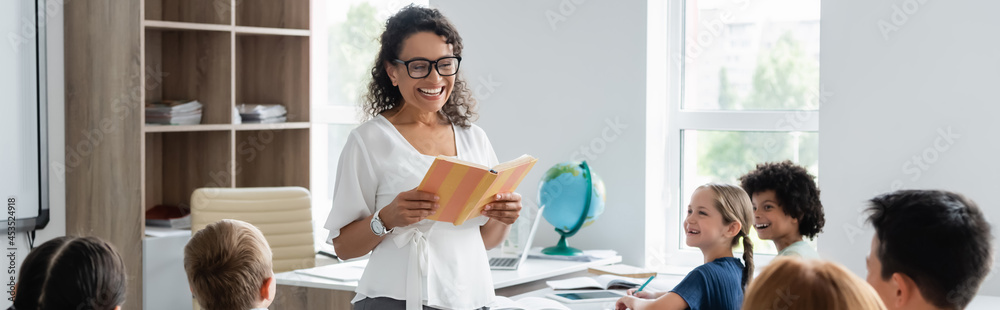 cheerful african american teacher with book smiling near multiethnic children, banner cheerful african american teacher with book smiling near multiethnic children, banner