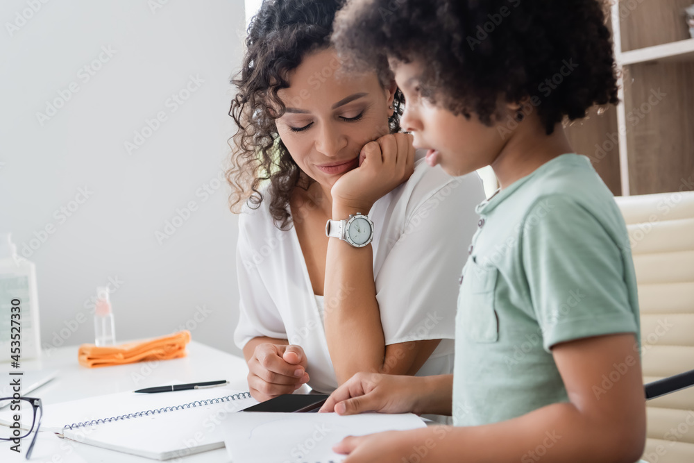 african american boy holding notebook near teacher in classroom african american boy holding notebook near teacher in classroom