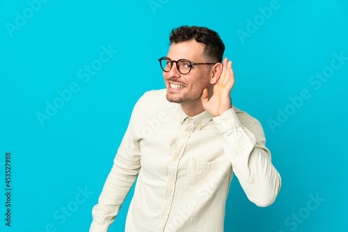 Young caucasian handsome man isolated on blue background listening to something by putting hand on the ear