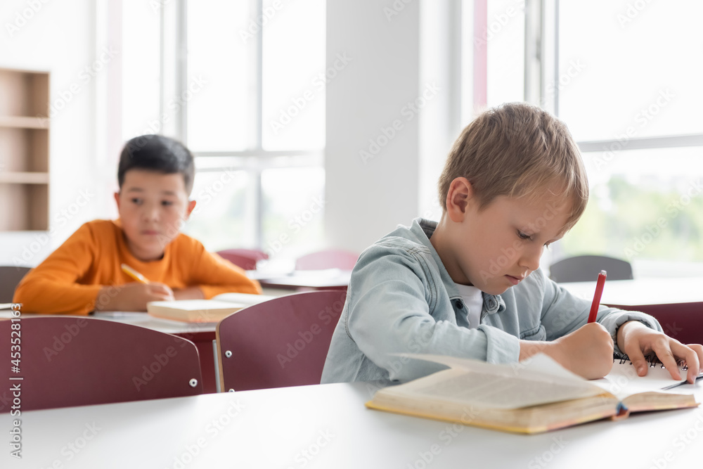 schoolboy writing during lesson near asian classmate on blurred background schoolboy writing during lesson near asian classmate on blurred background