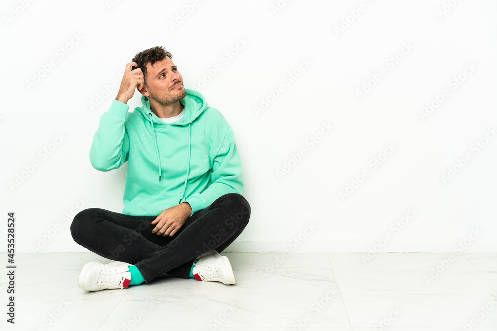 Young handsome caucasian man sitting on the floor having doubts and with confuse face expression Young handsome caucasian man sitting on the floor having doubts and with confuse face expression