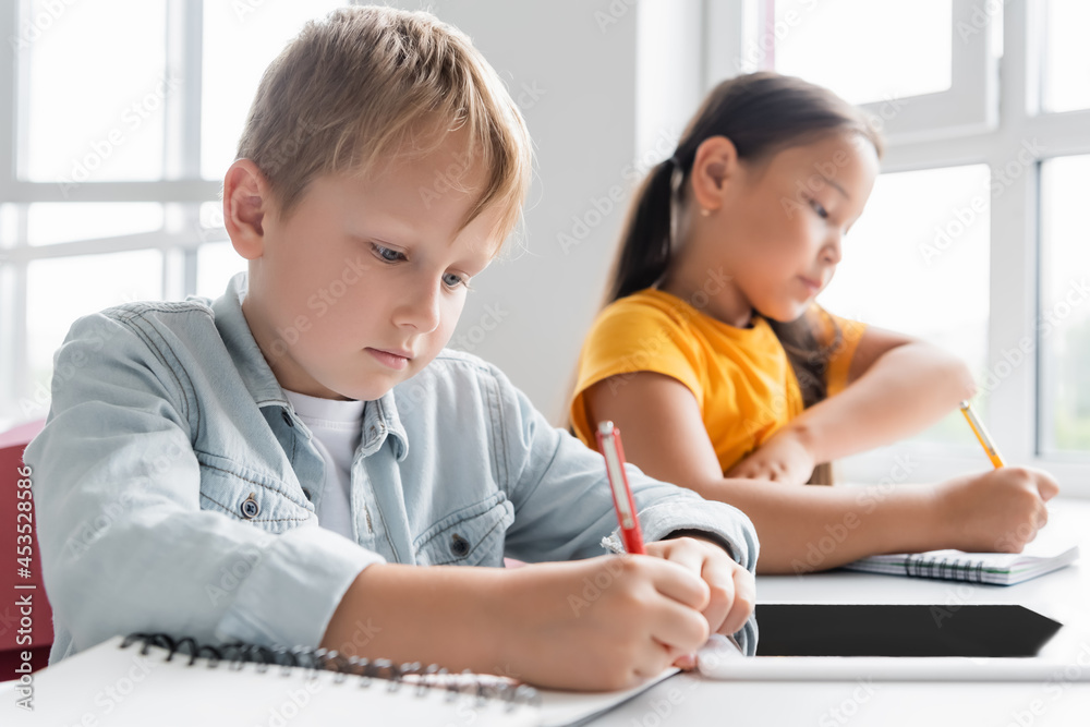 schoolboy writing in notebook near digital tablet and blurred asian girl schoolboy writing in notebook near digital tablet and blurred asian girl