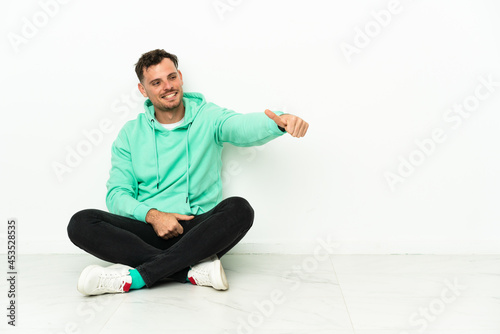 Young handsome caucasian man sitting on the floor giving a thumbs up gesture