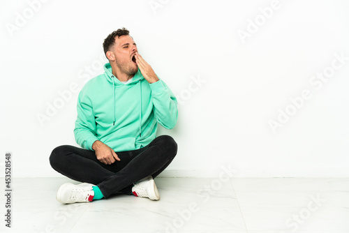 Young handsome caucasian man sitting on the floor yawning and covering wide open mouth with hand